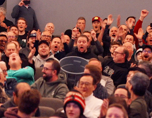 Many men cheer and yell as someone almost lands a roll of toilet paper in a garbage bin